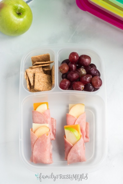 ham, cheese and apple wraps in a lunch box with crackers and grapes