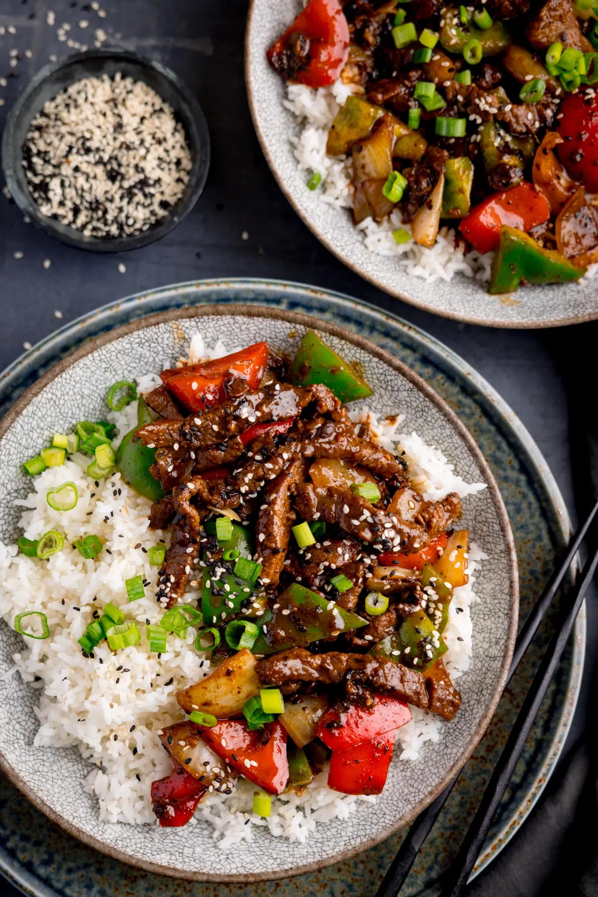 Beef in black bean sauce stir-fried with boiled rice in a bowl, topped with scallions and sesame seeds. The bowls are on a dark surface and there is another bowl on the plane. There is a small bowl of black and white sesame seeds nearby.