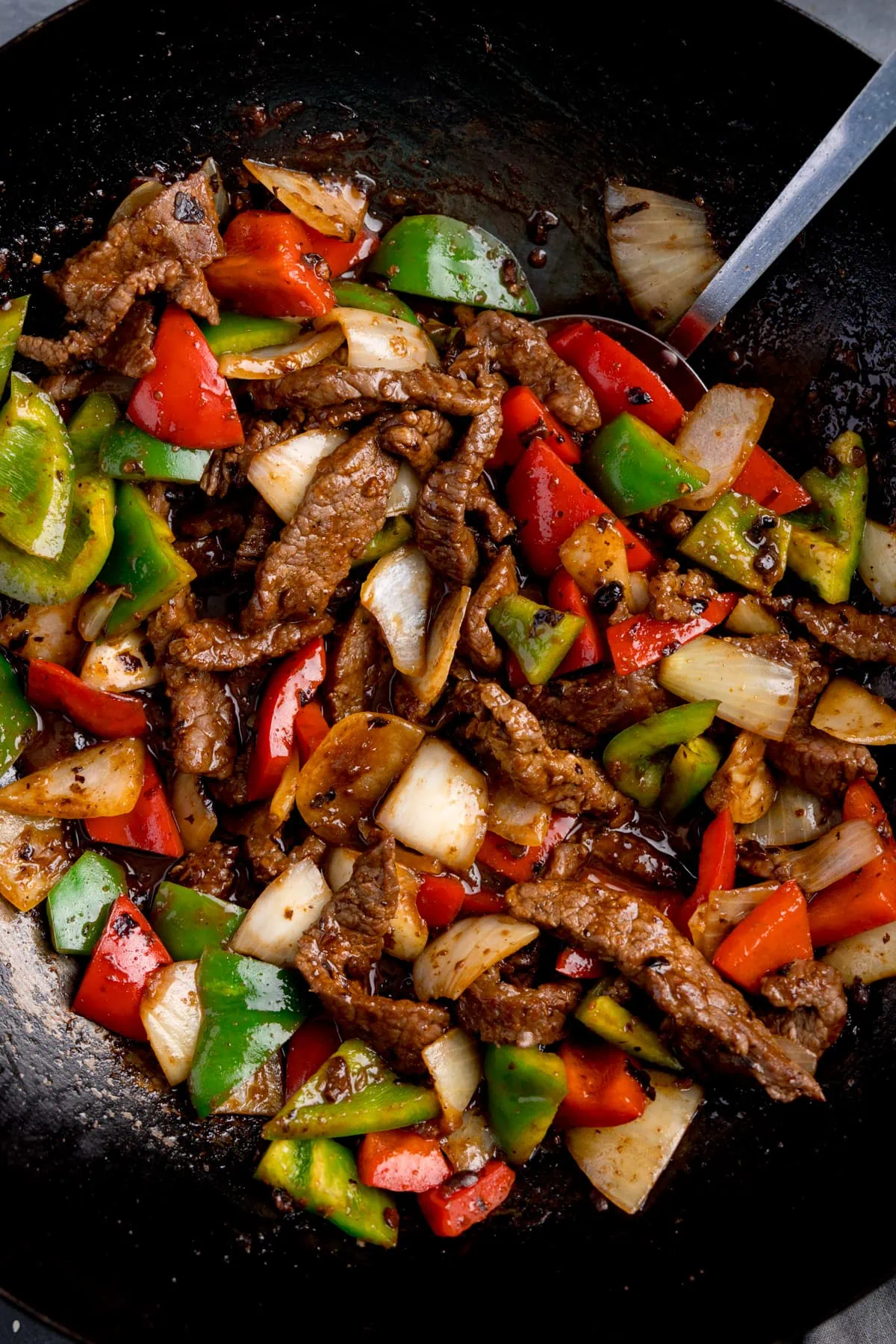 Close up of beef stir fry in black bean sauce, with bell peppers and onions, in a wok, with a spatula sticking out.
