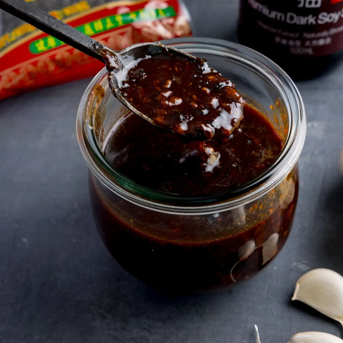 square image of a glass jar filled with homemade black bean sauce. There is a black spoon, taking a spoonful from the jar. The jar is on a dark surface and there are ingredients at the bottom.