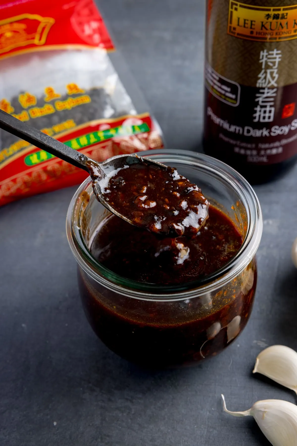 A glass jar filled with homemade black bean sauce. There is a black spoon, taking a spoonful from the jar. The jar is on a dark surface and there are scattered ingredients.