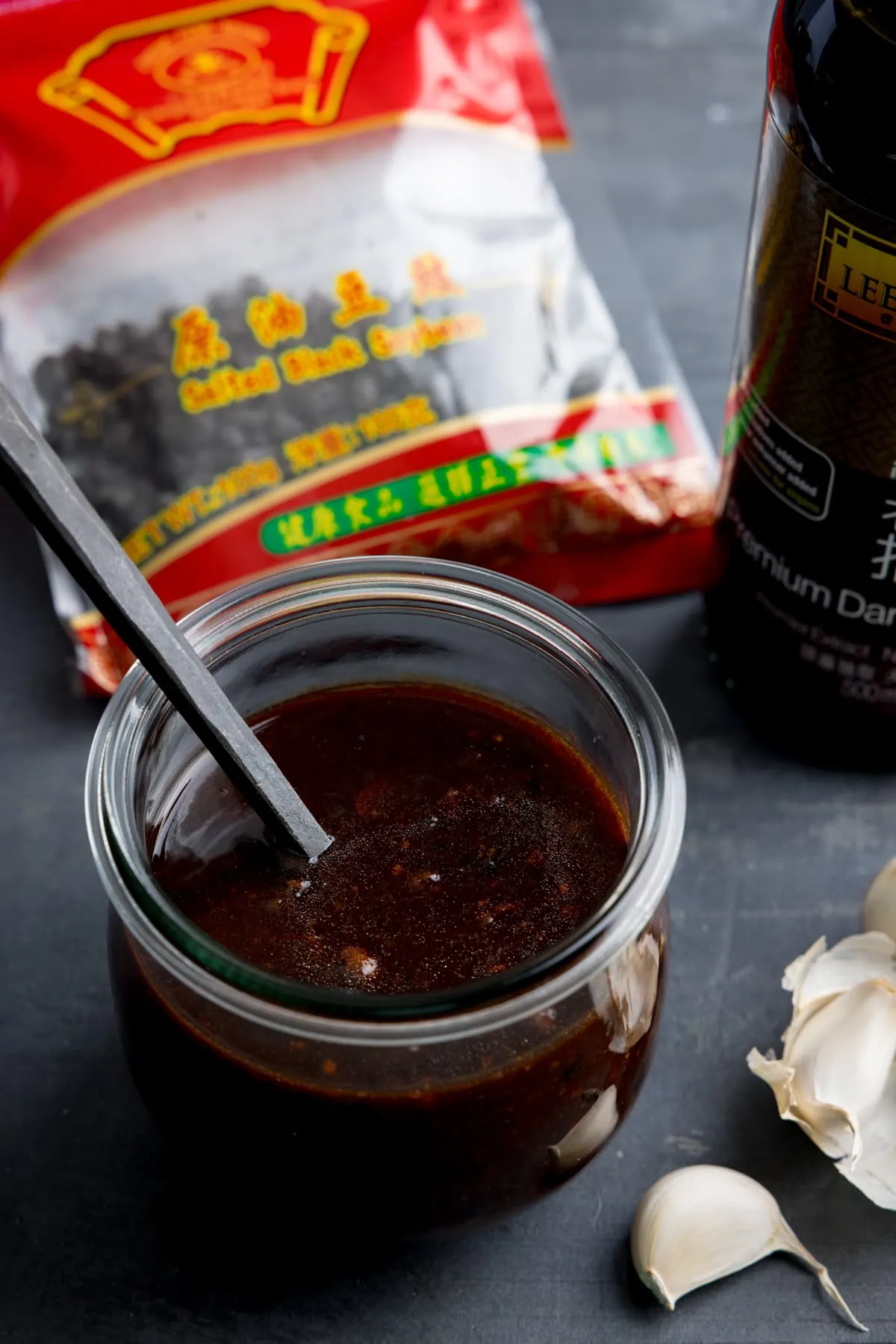 A glass jar filled with homemade black bean sauce. There is a black spoon sticking out of the jar. The jar is on a dark surface and there are ingredients at the bottom.