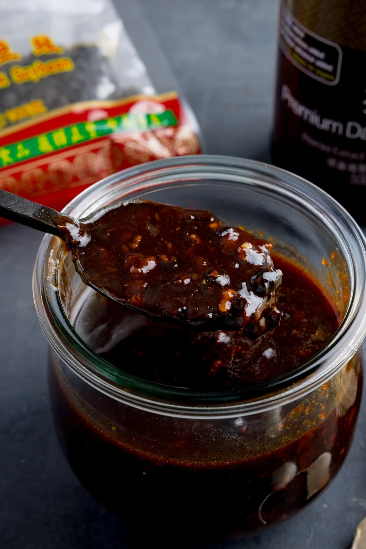 A glass jar filled with homemade black bean sauce. There is a black spoon, taking a spoonful from the jar. The jar is on a dark surface and there are ingredients at the bottom.