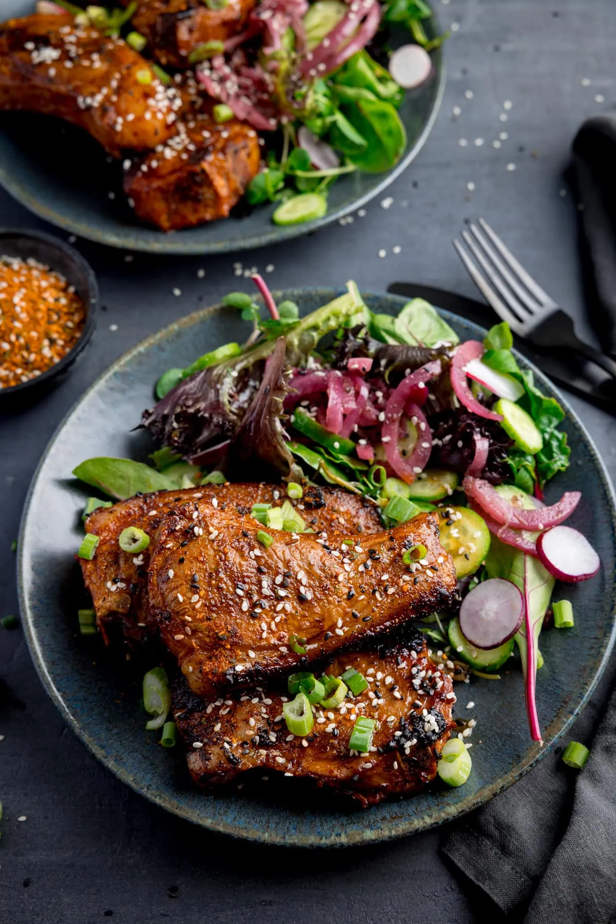 3 marinated and cooked lamb chops on a dark blue plate with a salad. The lamb chops are sprinkled with sesame seeds and chopped spring onions. The plate is on a dark gray background, next to a dark knife and fork and another plate of lamb chops.