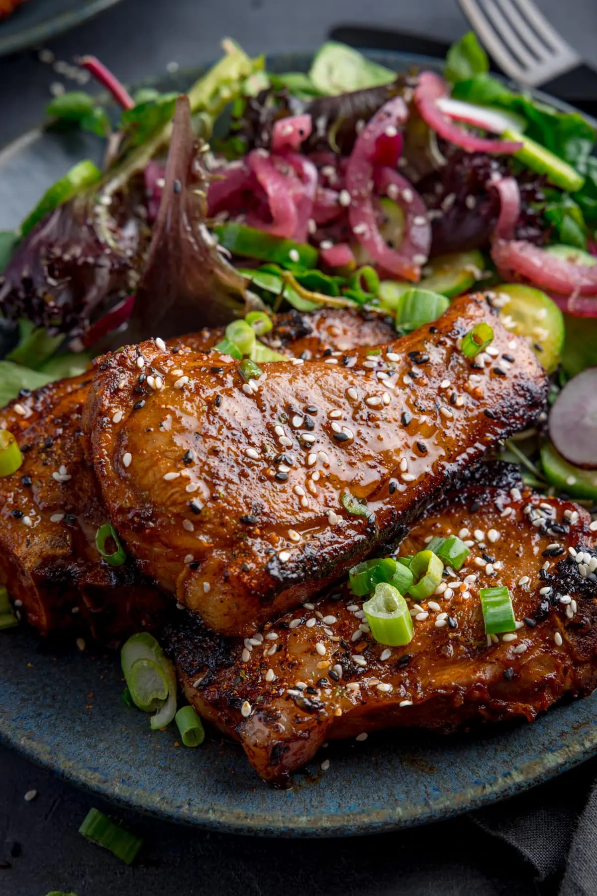 3 marinated and cooked lamb chops on a dark blue plate with a salad. The lamb chops are sprinkled with sesame seeds and chopped spring onions.