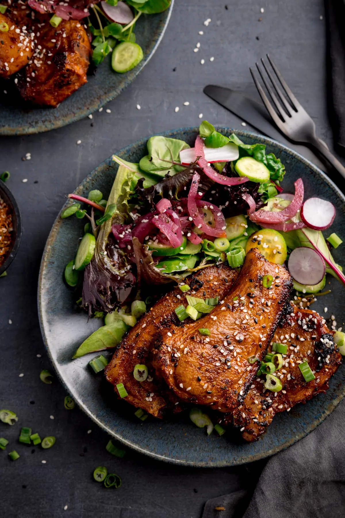 Top image of 3 marinated and cooked lamb chops on a dark blue plate next to a salad. The lamb chops are sprinkled with sesame seeds and chopped spring onions. The plate is on a dark gray background, next to a dark knife and fork and another plate of lamb chops.