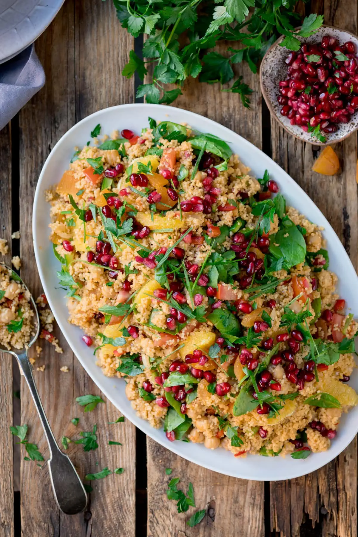 Top image of a large oval white plate filled with Moroccan-style couscous with oranges and pomegranates. The plate is on a wooden table, next to a spoonful of couscous, a plate of pomegranate, and fresh herbs scattered throughout.