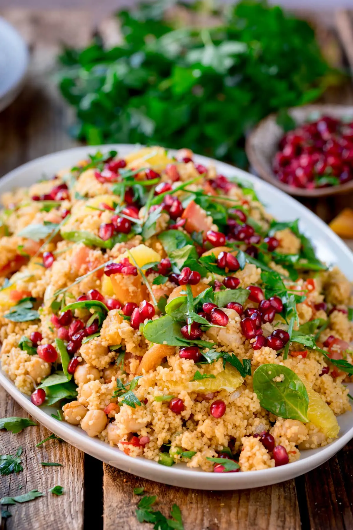 Side image of a large oval serving dish filled with Moroccan-style couscous, topped with herbs and pomegranate. The plate is on a wooden table and there are fresh herbs and a small bowl of pomegranate in the background.