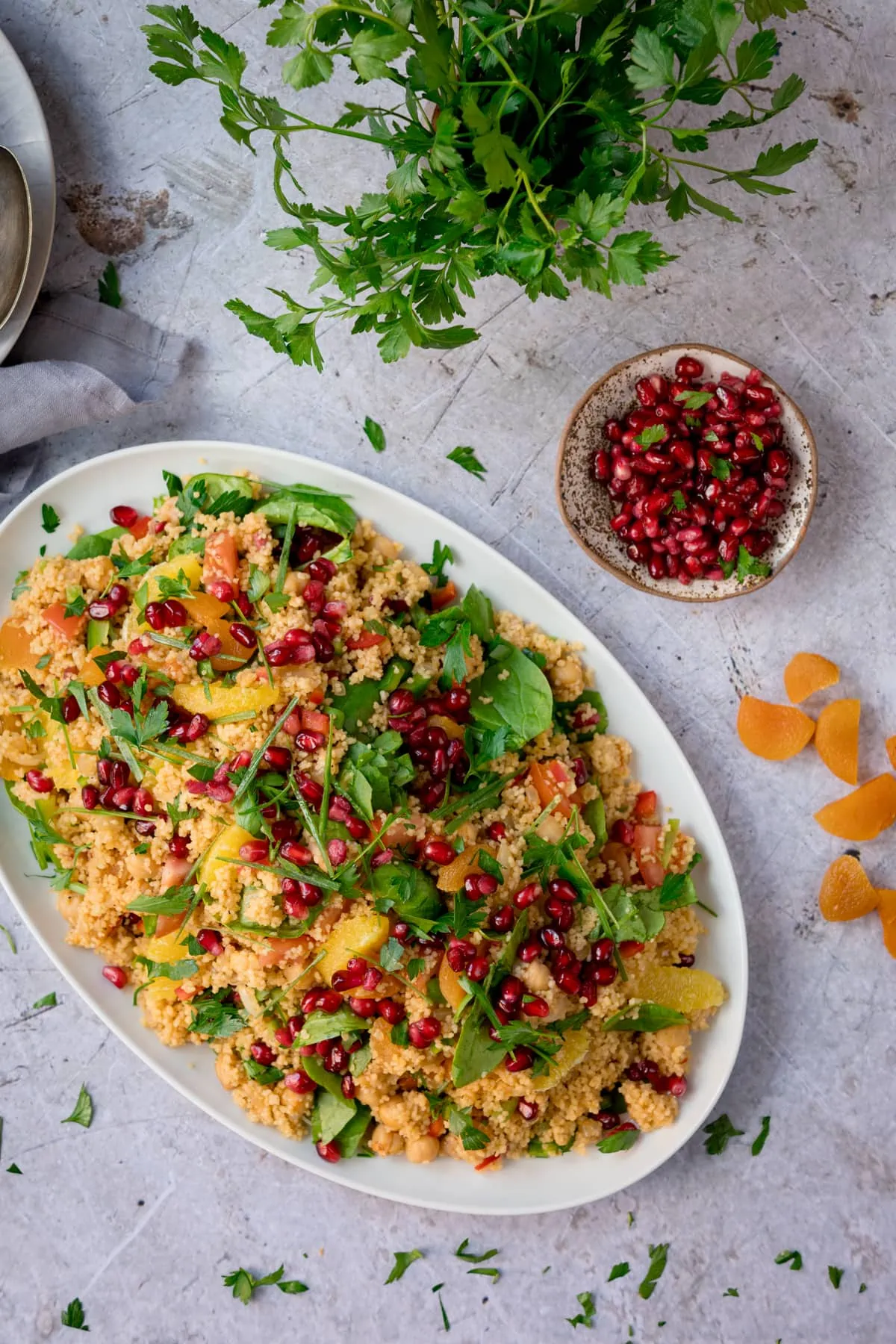 Top image of a large oval white plate filled with Moroccan-style couscous with oranges and pomegranates. The plate is on a white surface next to a plate of pomegranate. There are chopped dried apricots and fresh herbs scattered throughout.