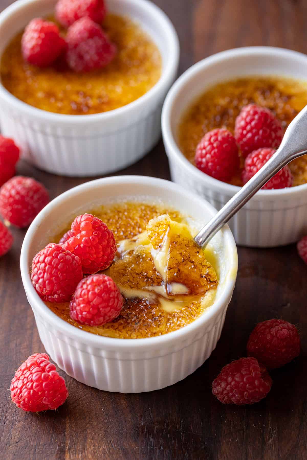 Three ramekins of creme brulee topped with raspberries, one with the caramelized sugar topping cracked with a spoon.