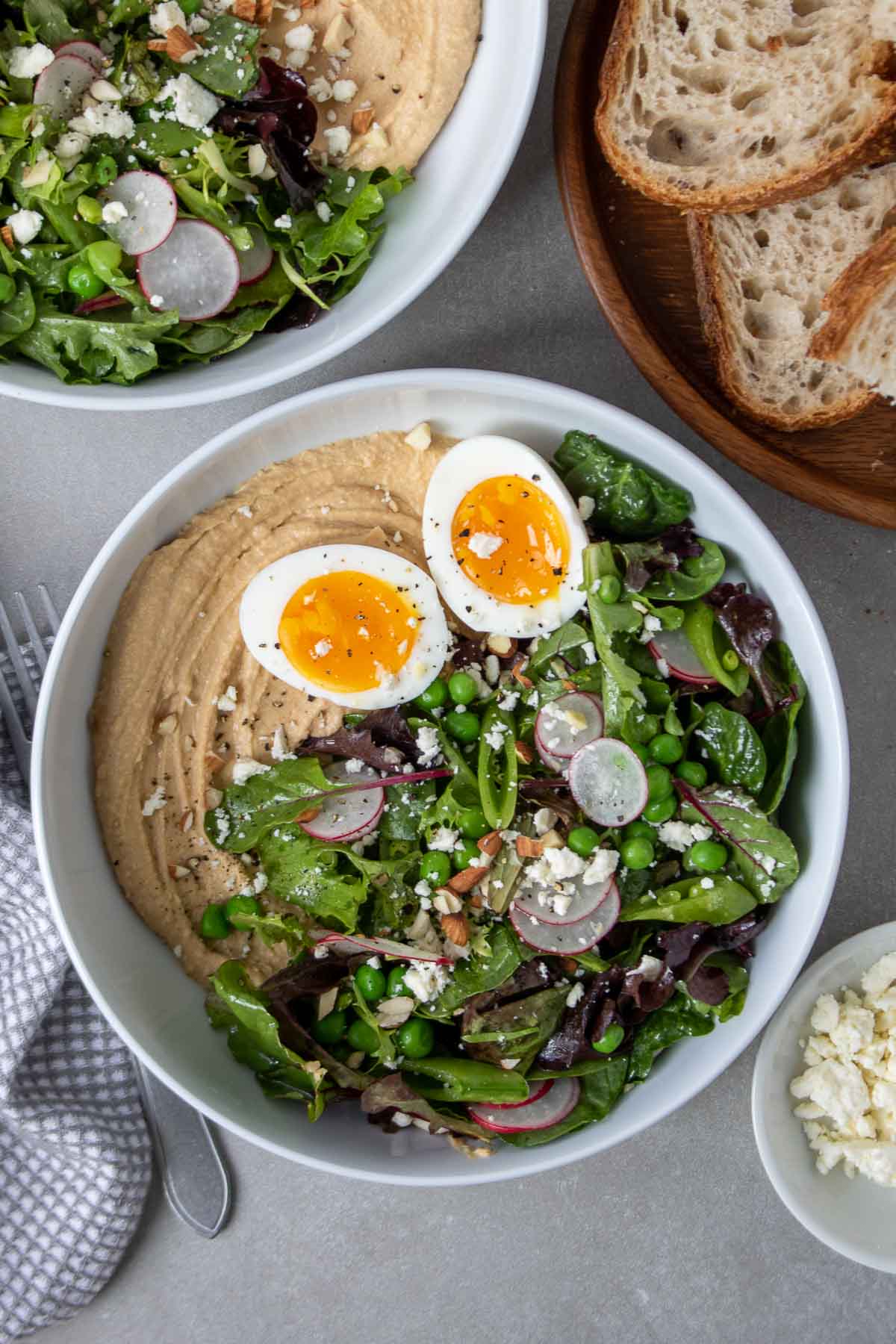 Overhead image of bowl with spread of hummus, topped with salad, a jammy 8-minute egg, feta cheese, and chopped almonds, salt, and pepper, with a side of sourdough bread.