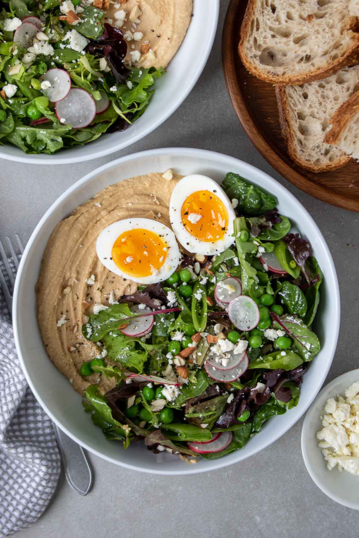 Vue d'en haut d'un bol avec du houmous, garni de salade, d'un œuf à la coque de 8 minutes, de fromage feta émietté et d'amandes hachées, assaisonné de sel et de poivre, avec un morceau de pain de campagne en accompagnement.