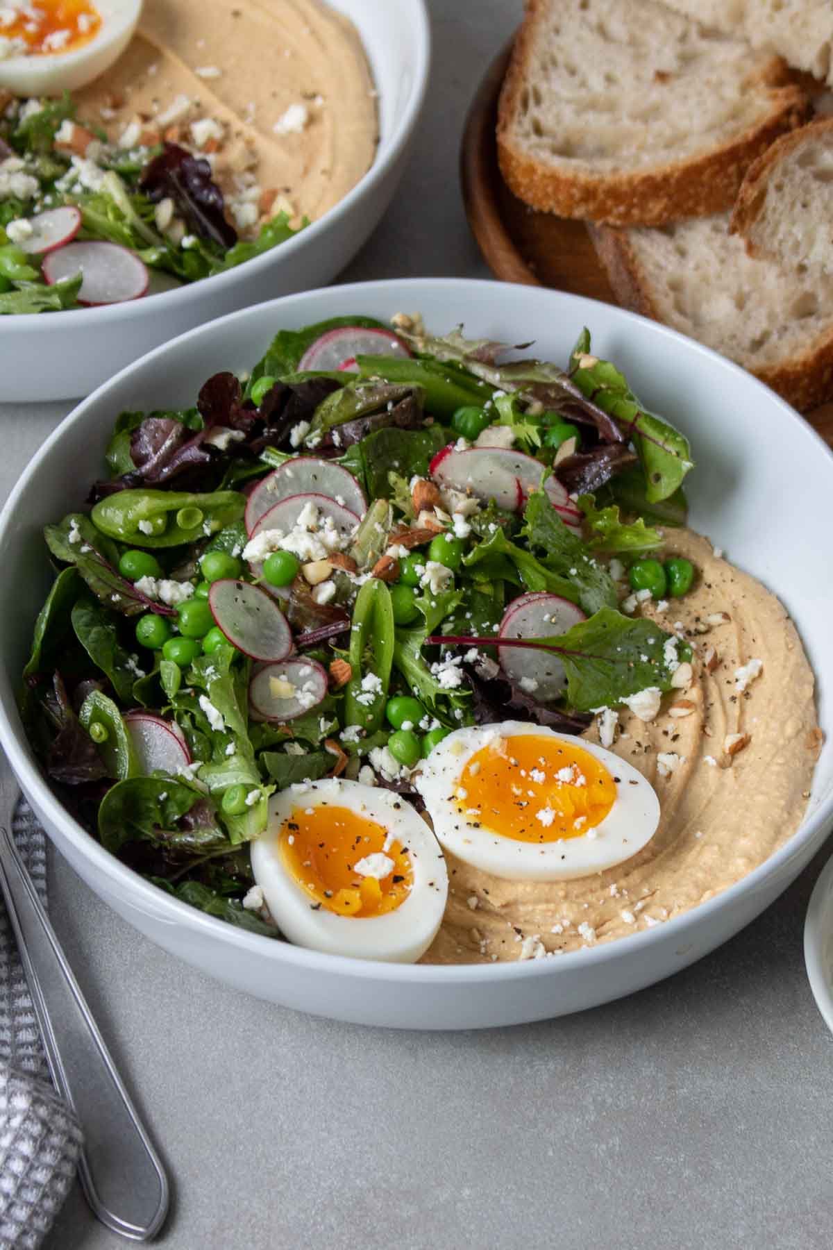 Two bowls filled with hummus and salad, topped with a soft-boiled egg, feta cheese, chopped almonds, and a side of sourdough bread.