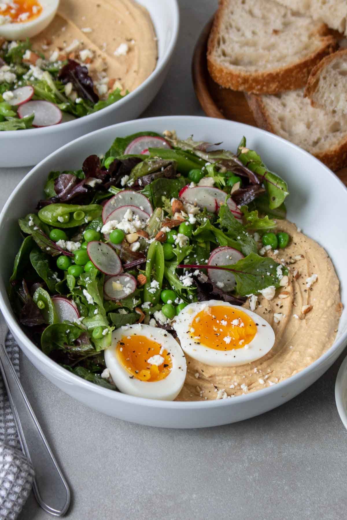 Deux bols remplis de houmous et de salade, garnis d'un œuf à la coque, de fromage feta, d'amandes hachées et d'un morceau de pain de campagne.