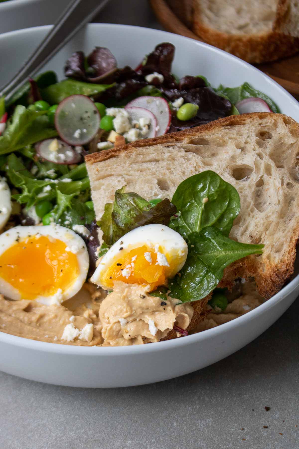 Close up of slice of bread in a bowl with some leafy greens, hummus, and a piece of boiled egg on the bread.