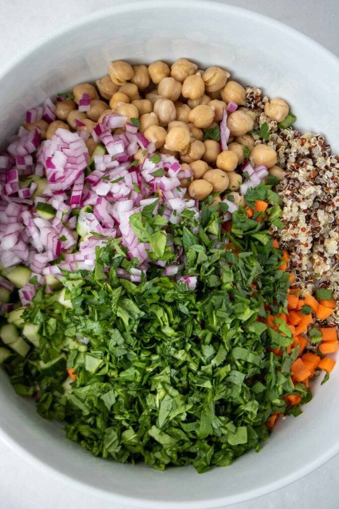 Unmixed salad ingredients in a large white bowl: tri-color quinoa, chickpeas, red onion, cucumber, carrots, spinach, and parsley.