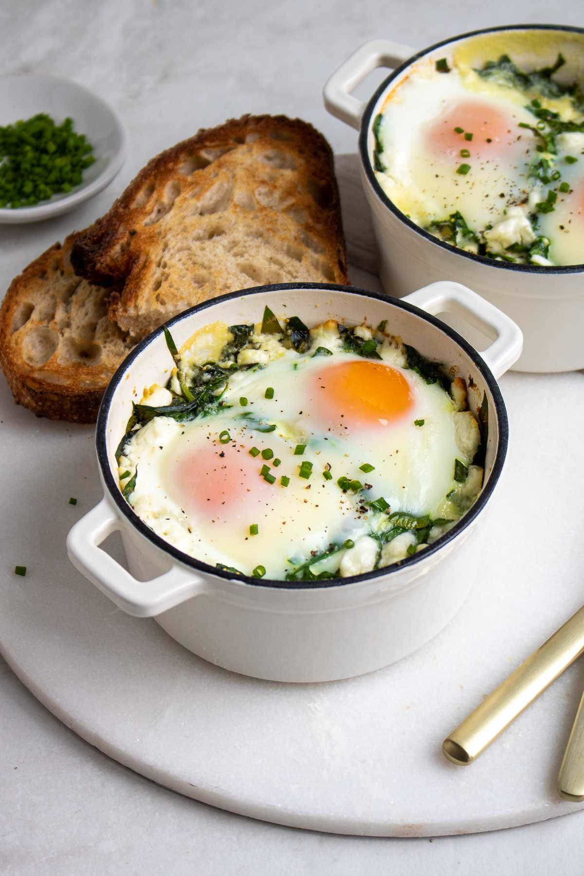 Two ramekins filled with baked eggs with spinach and goat cheese with a side of toast.