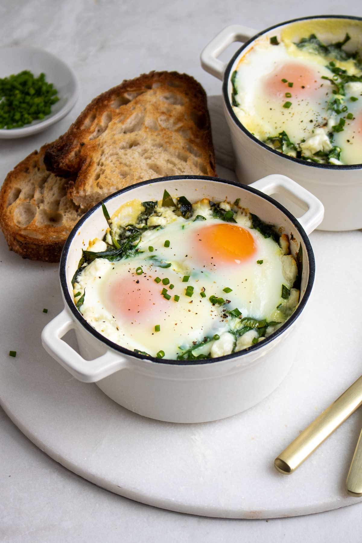 Deux ramequins remplis d'œufs au four aux épinards et au fromage de chèvre avec une tranche de pain grillé.