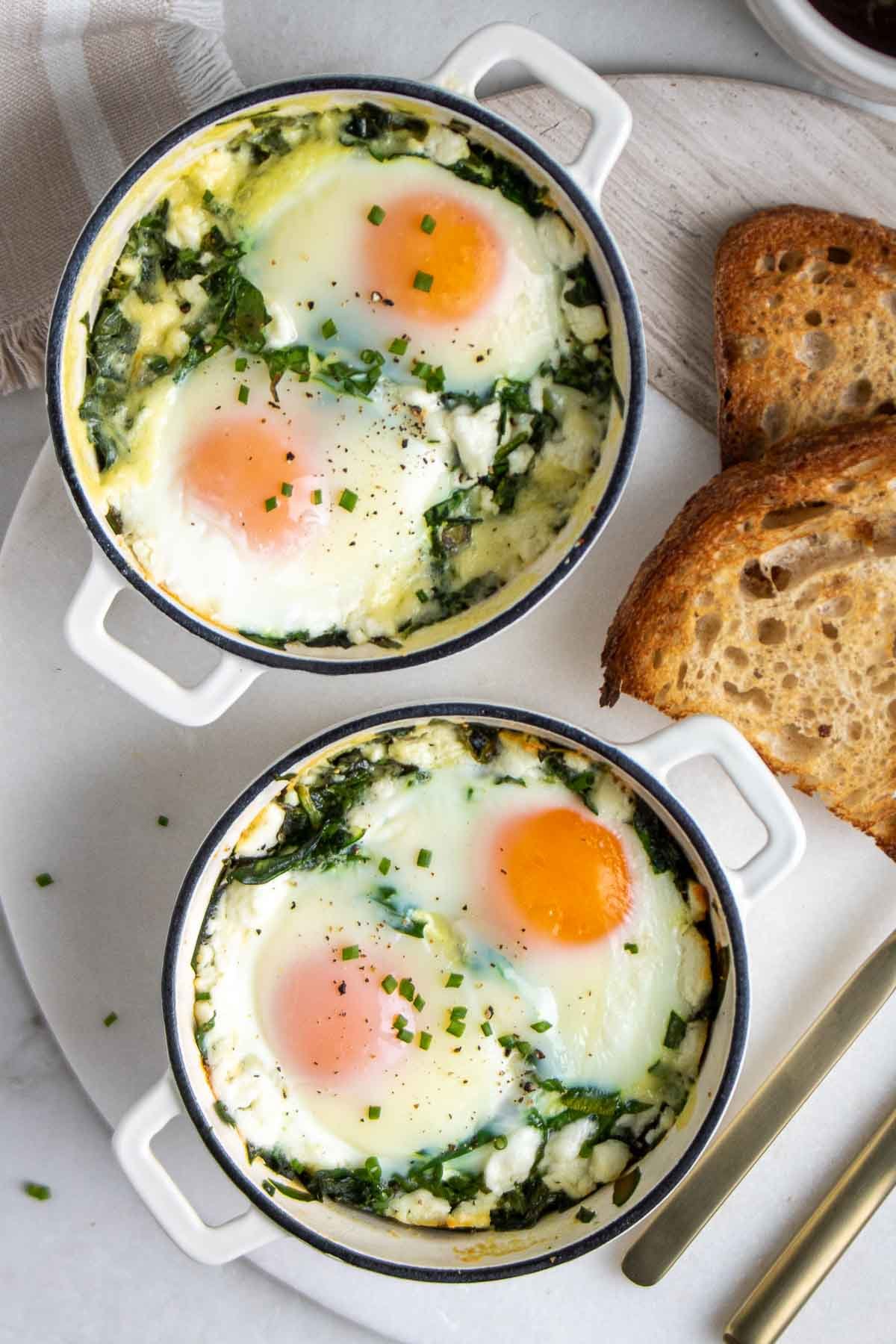 Two ramekins of creamy spinach baked eggs on a white background with a side of toast and spinach.