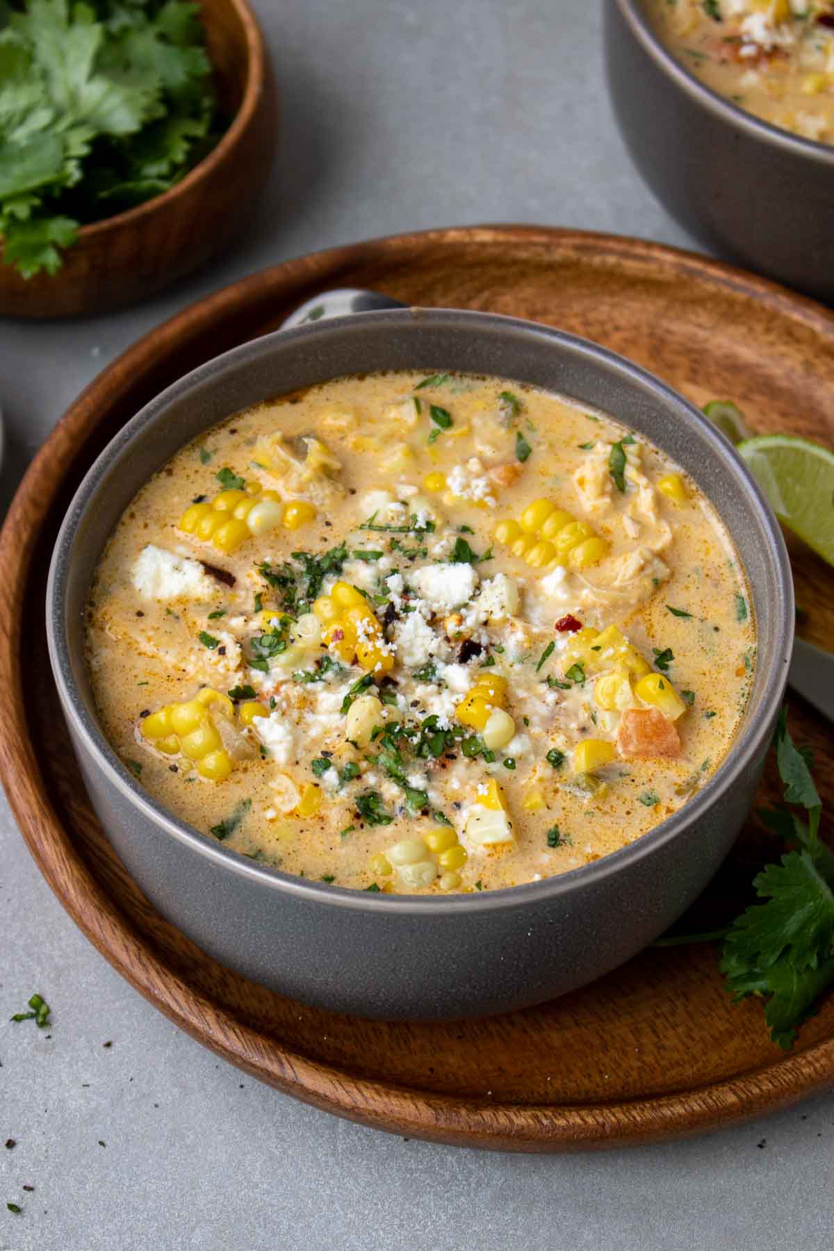 Corn chicken soup in a grey bowl on a wood plate with cilantro and cotija cheese on top.