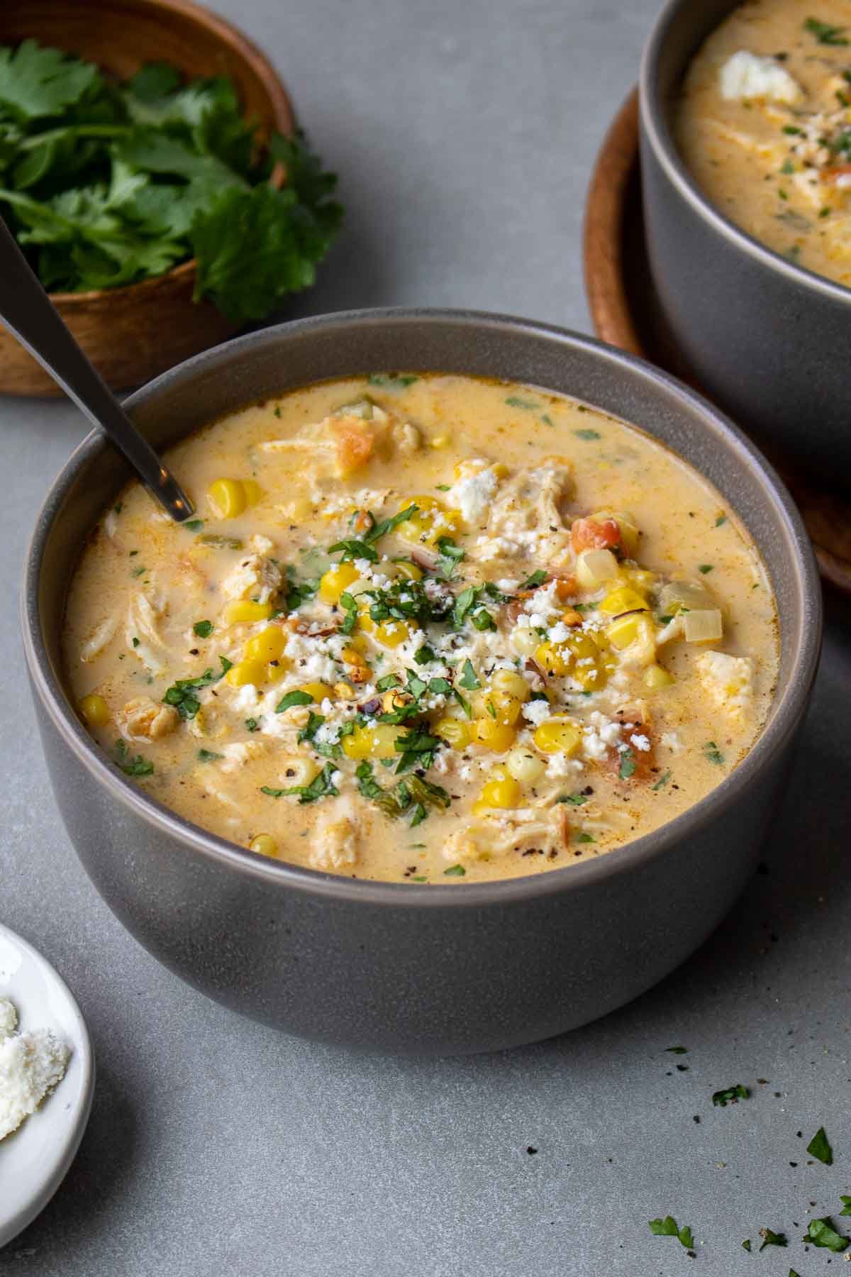 Two bowls of chicken corn soup in grey bowls with a spoon on a dark grey background.