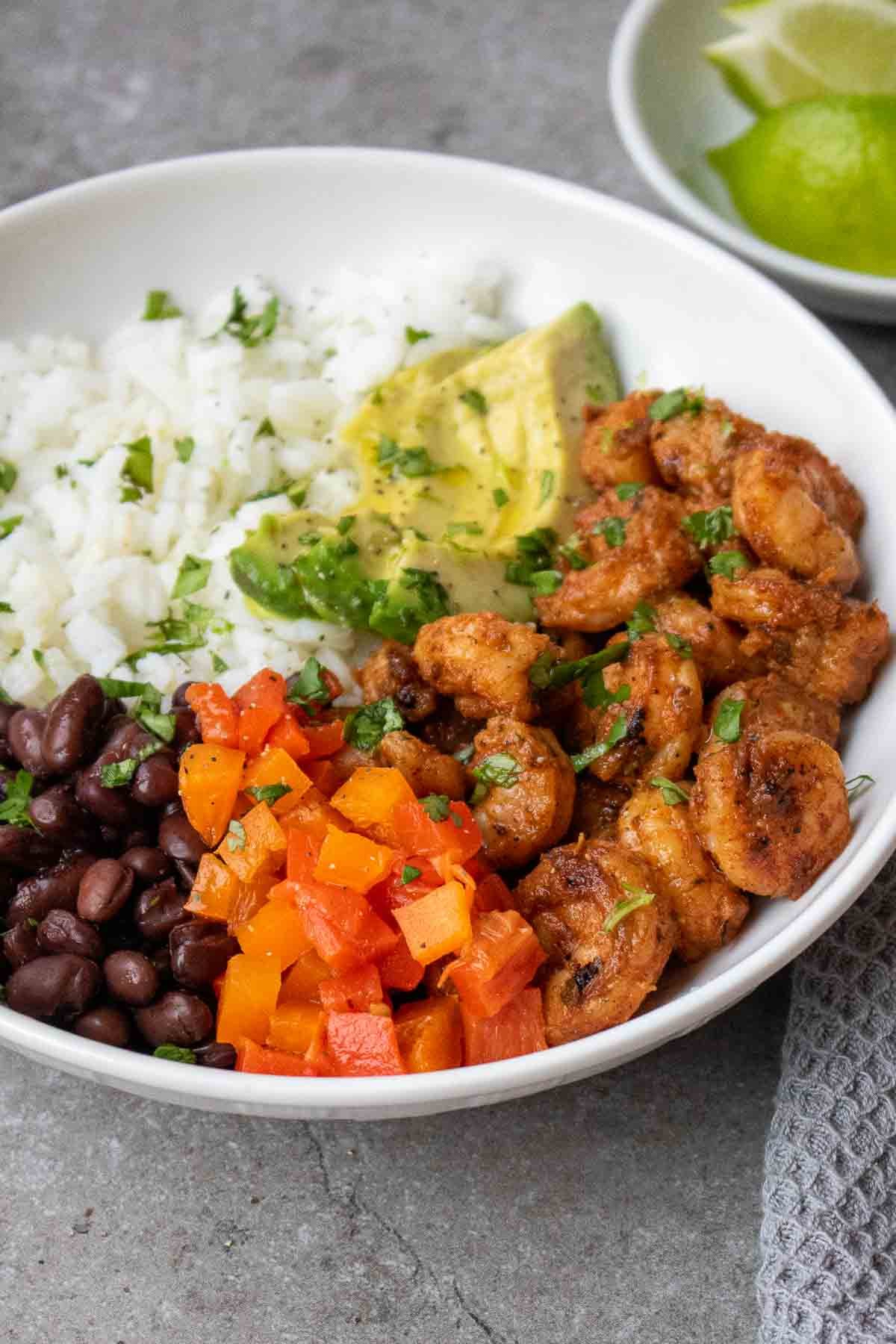 Bowl with rice, black beans, bell peppers, avocado and blackened shrimp with cilantro and lime wedges.