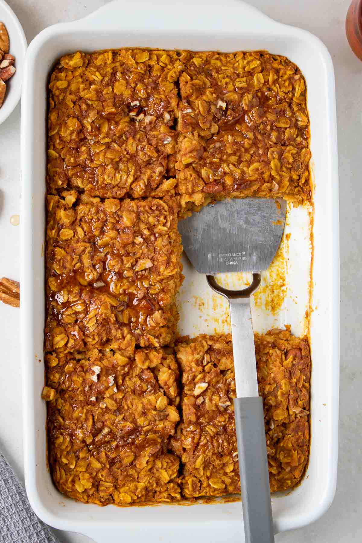 Pumpkin baked oatmeal in a white baking dish sliced into 6 pieces with maple syrup on top and a spatula on a white background