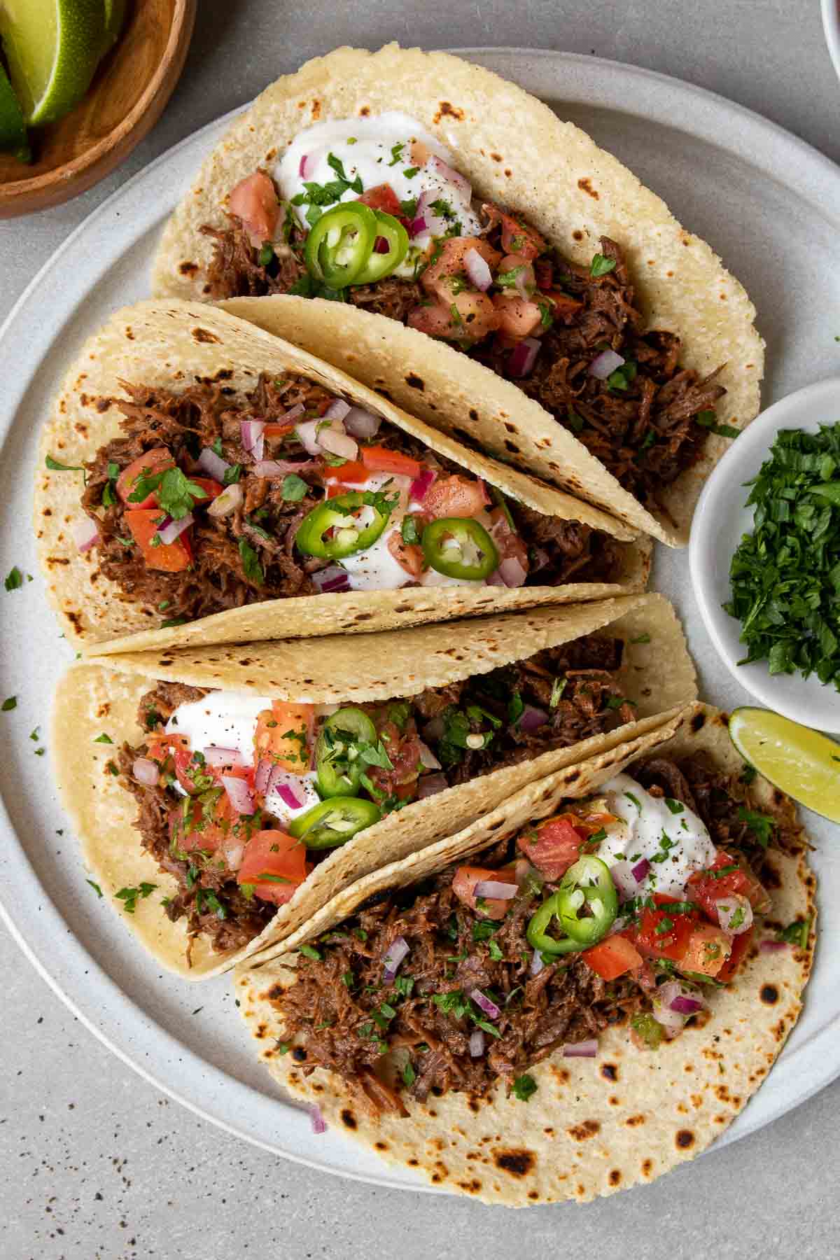 Overhead shot of four slow-cooker shredded beef tacos on a plate with toppings and a side of lime and cilantro.
