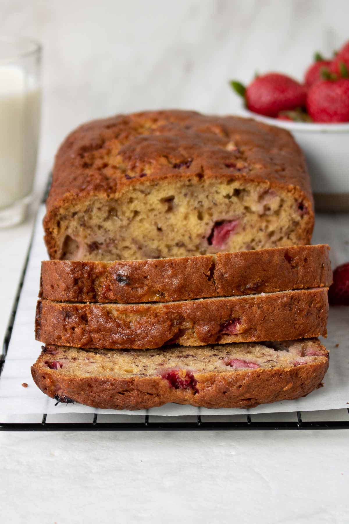 Image of a strawberry banana bread loaf with three slices cut, placed on a baking rack alongside a glass of milk and a bowl of strawberries.