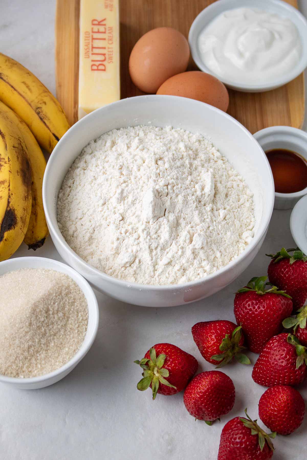 Image of the ingredients for strawberry banana bread, including flour, sugar, baking soda, salt, bananas, eggs, vanilla extract, butter, and fresh strawberries.