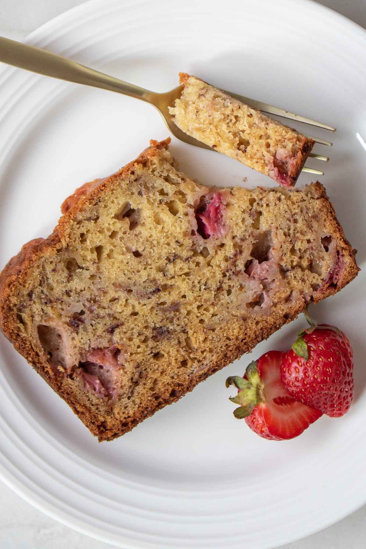 Image of a slice of strawberry banana bread on a white plate with a bite taken out with a fork.