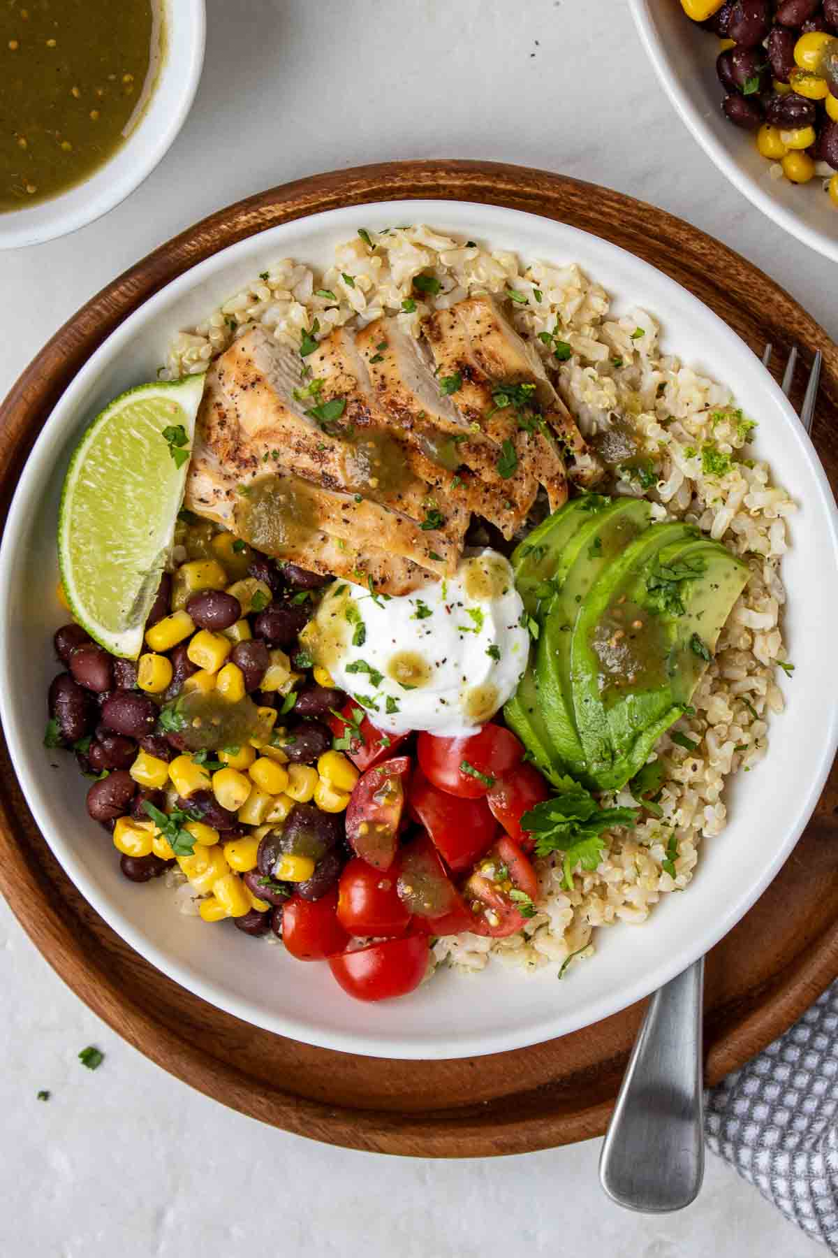 Baja bowl with chicken in a white bowl on a wood plate with a fork and a bowl of black bean and corn salsa and bowl of salsa verde on the side.