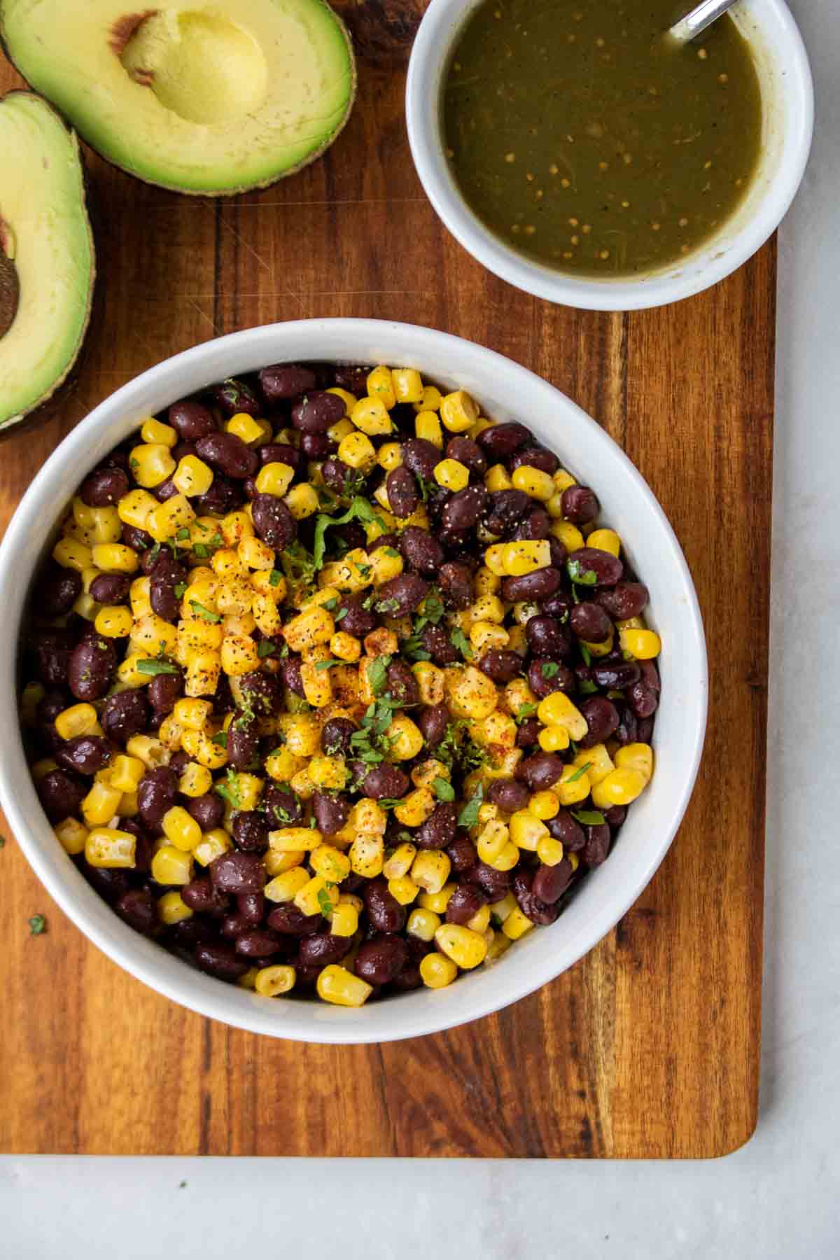 White bowl of black bean and corn salsa on a wood cutting board with an avocado and salsa verde on the side.