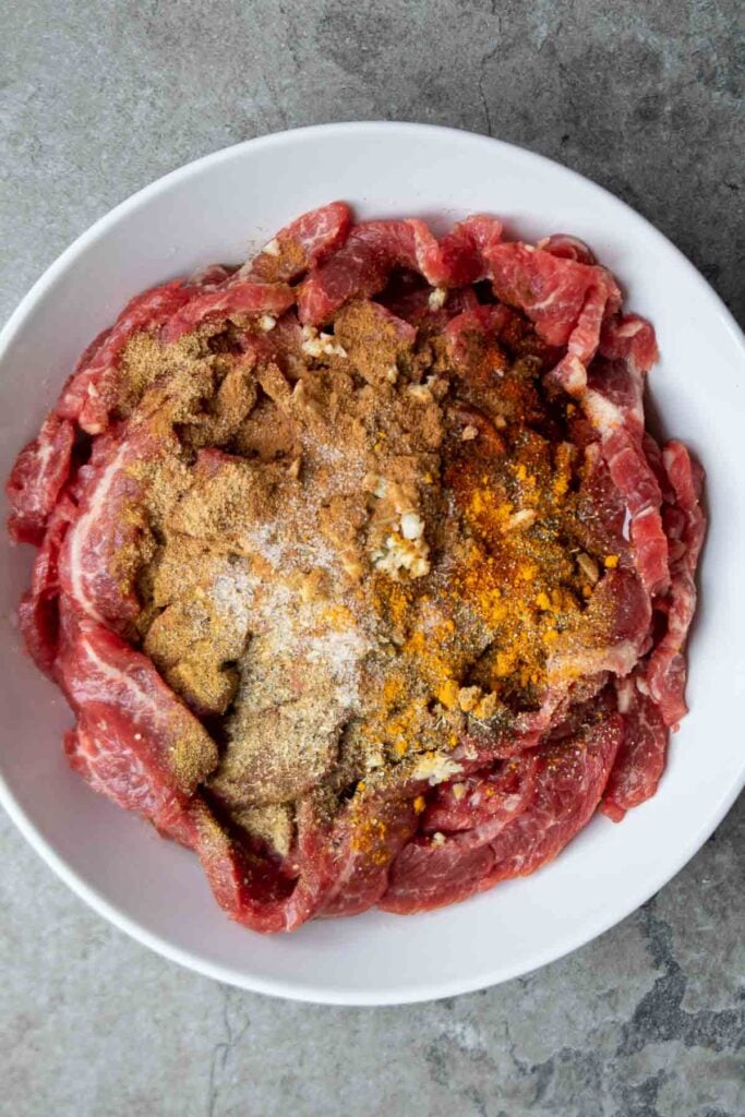 Thinly sliced flank steak in a white bowl with olive oil, lemon juice, minced garlic, and shawarma spice mix on top.