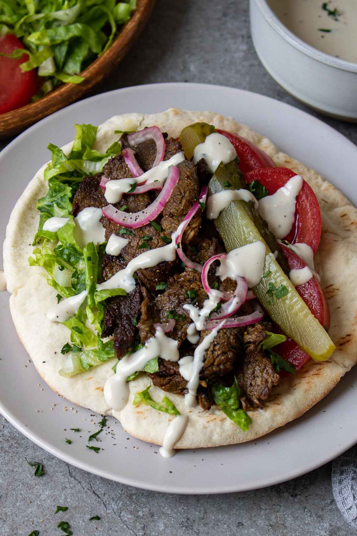 Plate with a pita layered with beef shawarma, shredded lettuce, slices of tomato, pickled red onion, pickle slices, and a drizzle of tahini sauce on top.
