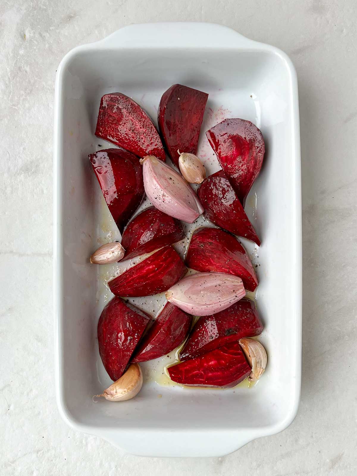 Raw beets, shallot, and garlic in olive oil in a white baking dish.