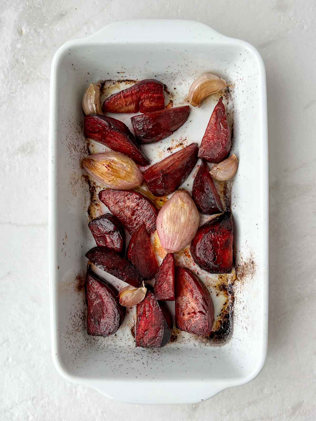 Roasted beets, shallot, and garlic in olive oil in a white baking dish.