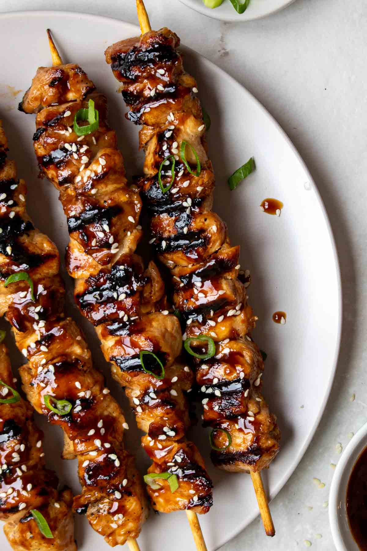 Grilled teriyaki chicken skewers on a plate with sesame seeds and green onion on top.
