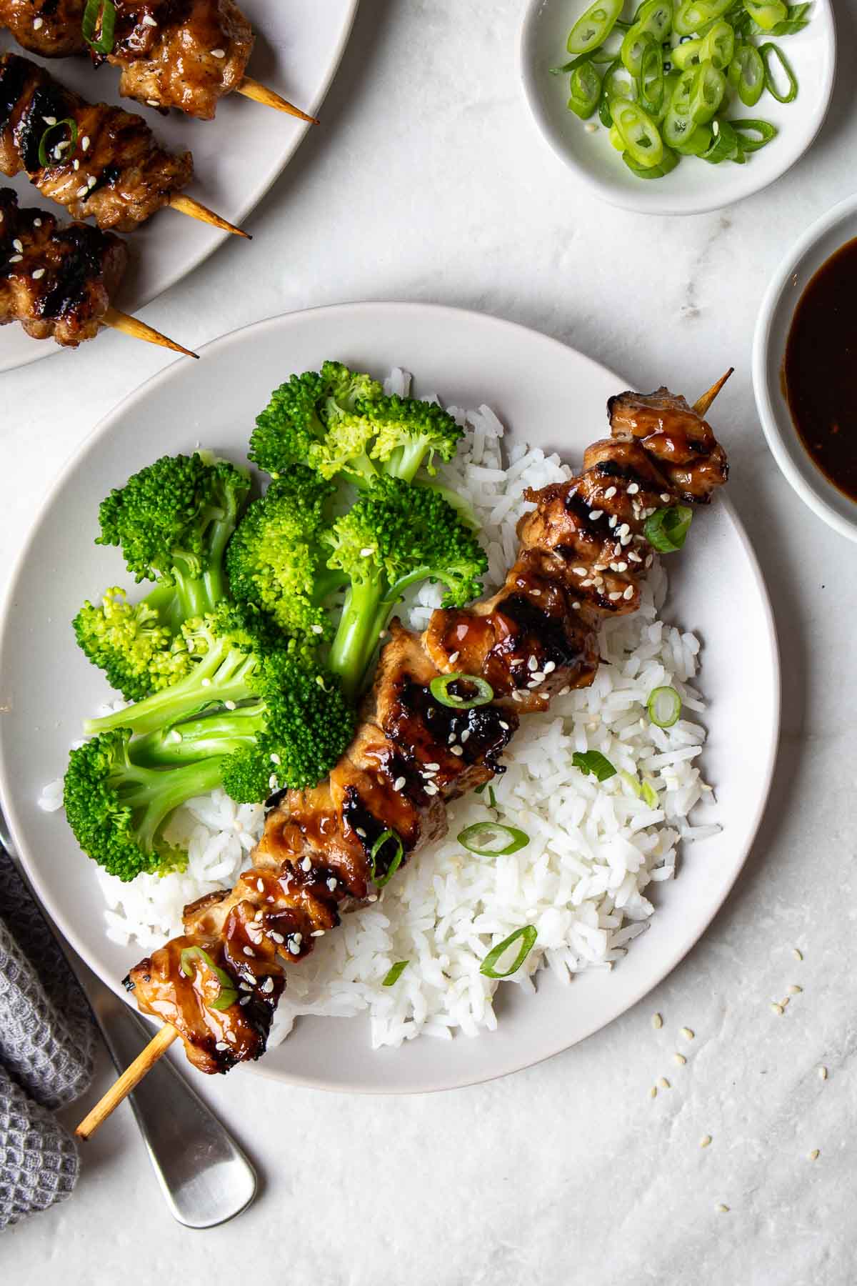 A plate of grilled teriyaki chicken skewers with a serving on a plate with rice, broccoli, sesame seeds, and green onion.