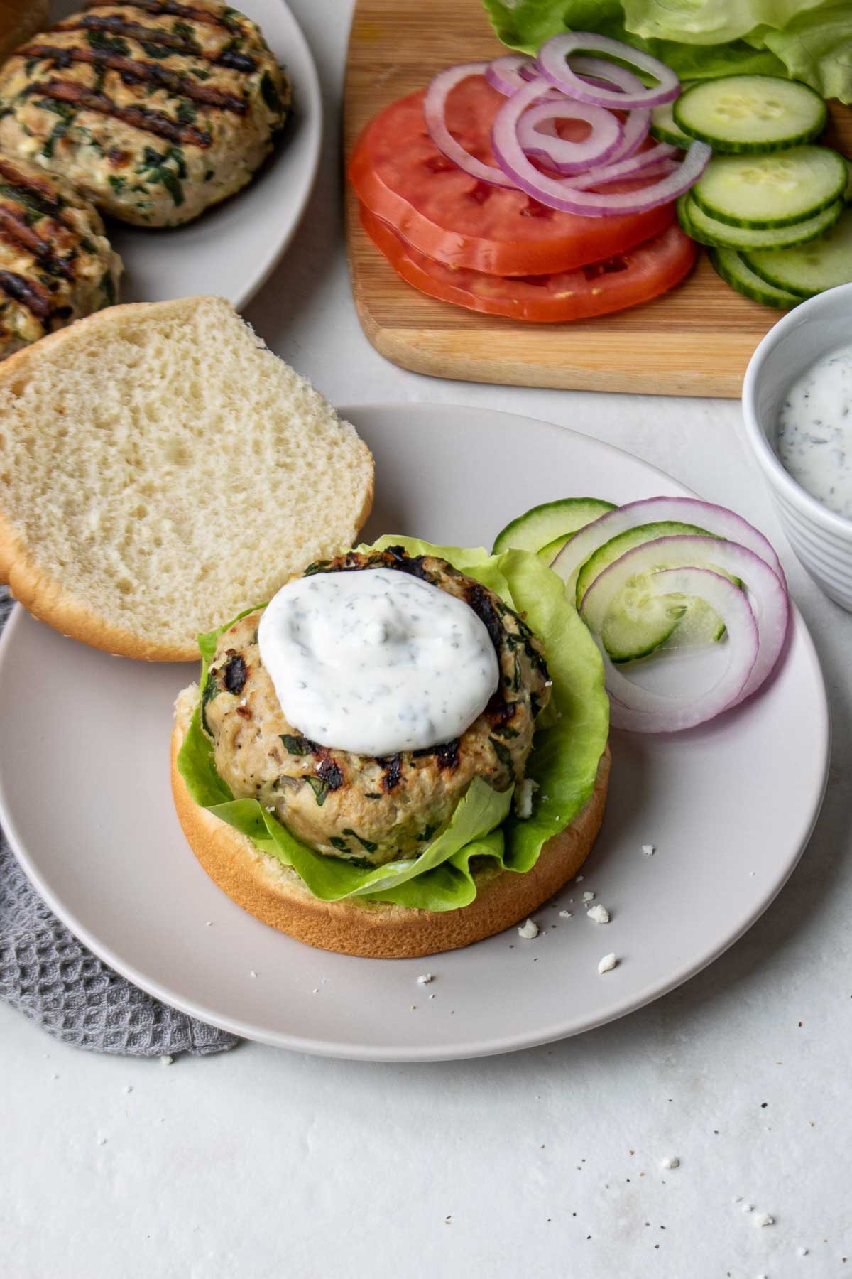 Cooked turkey burgers with spinach and feta being prepared with toppings on a cutting board.