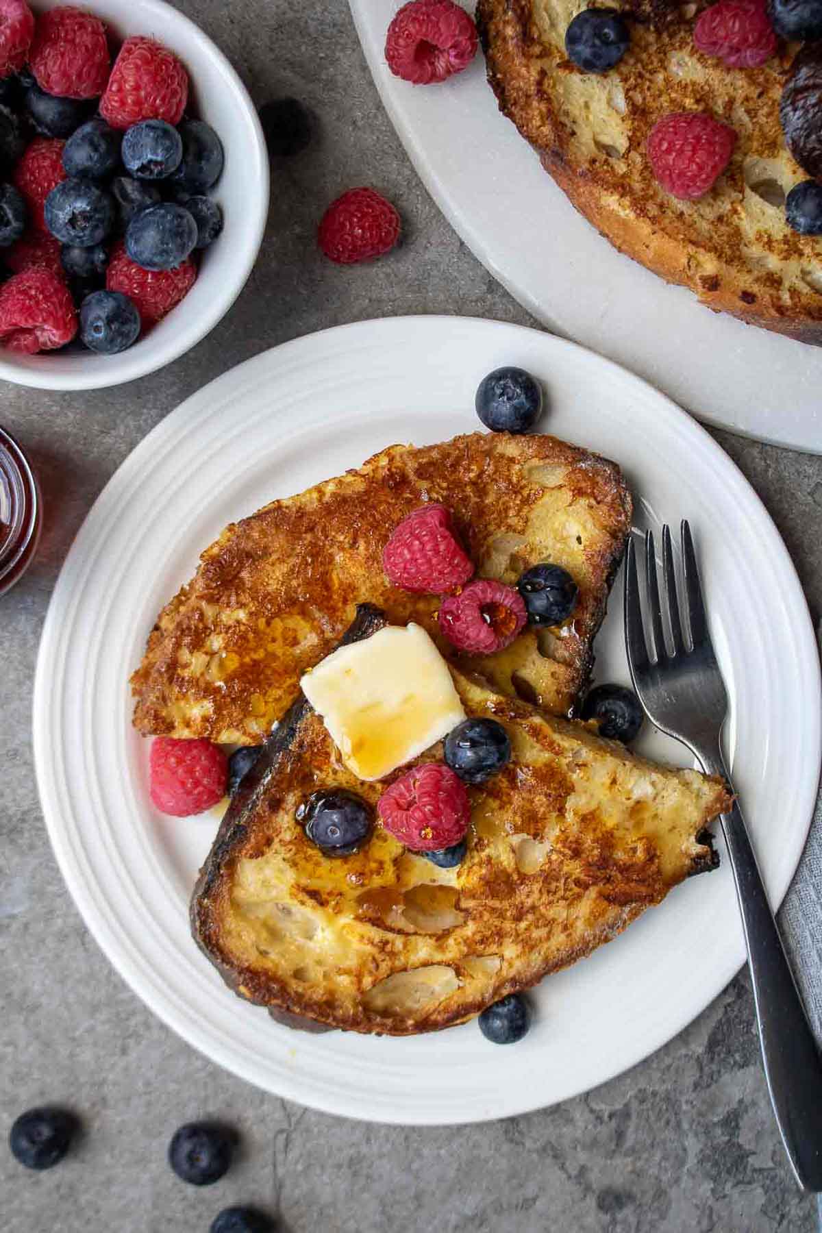 Two slices of sourdough French toast on a white plate with butter, raspberries, blueberries, and maple syrup. 