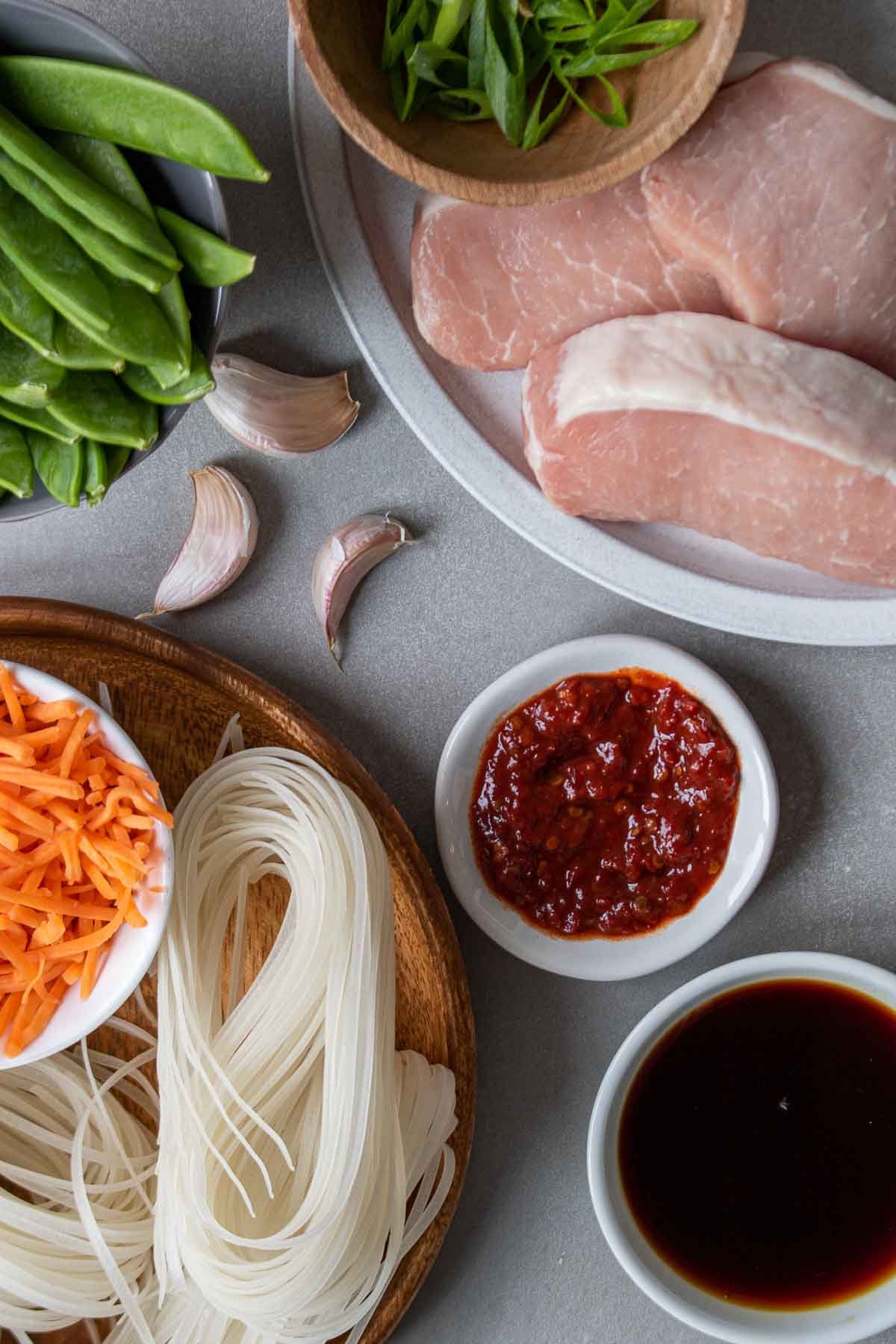 Ingredients for pork noodles; pork, rice noodles, chili paste, soy sauce, garlic, snow peas, and carrots.