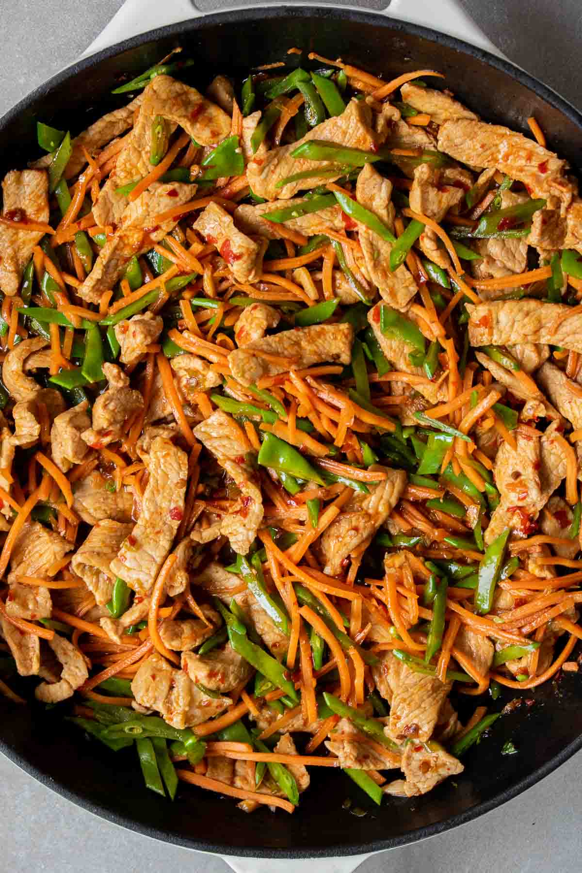 Cooked sliced pork with carrots and snow peas in a pan covered in a spicy sauce