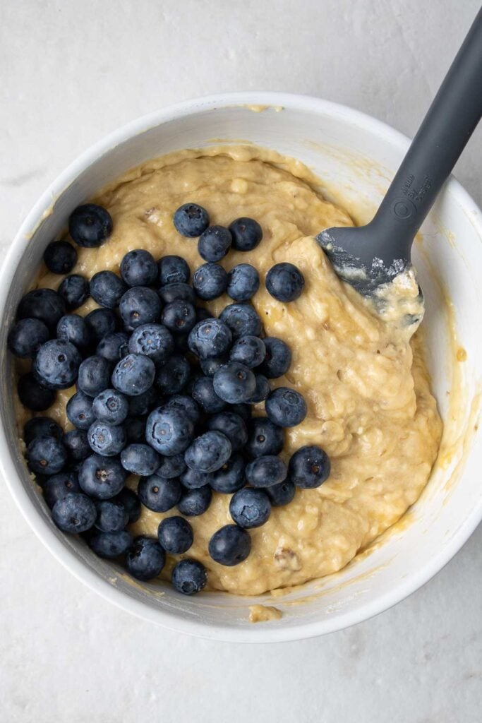 Mixed banana muffin batter in a bowl with fresh blueberries about to be mixed in