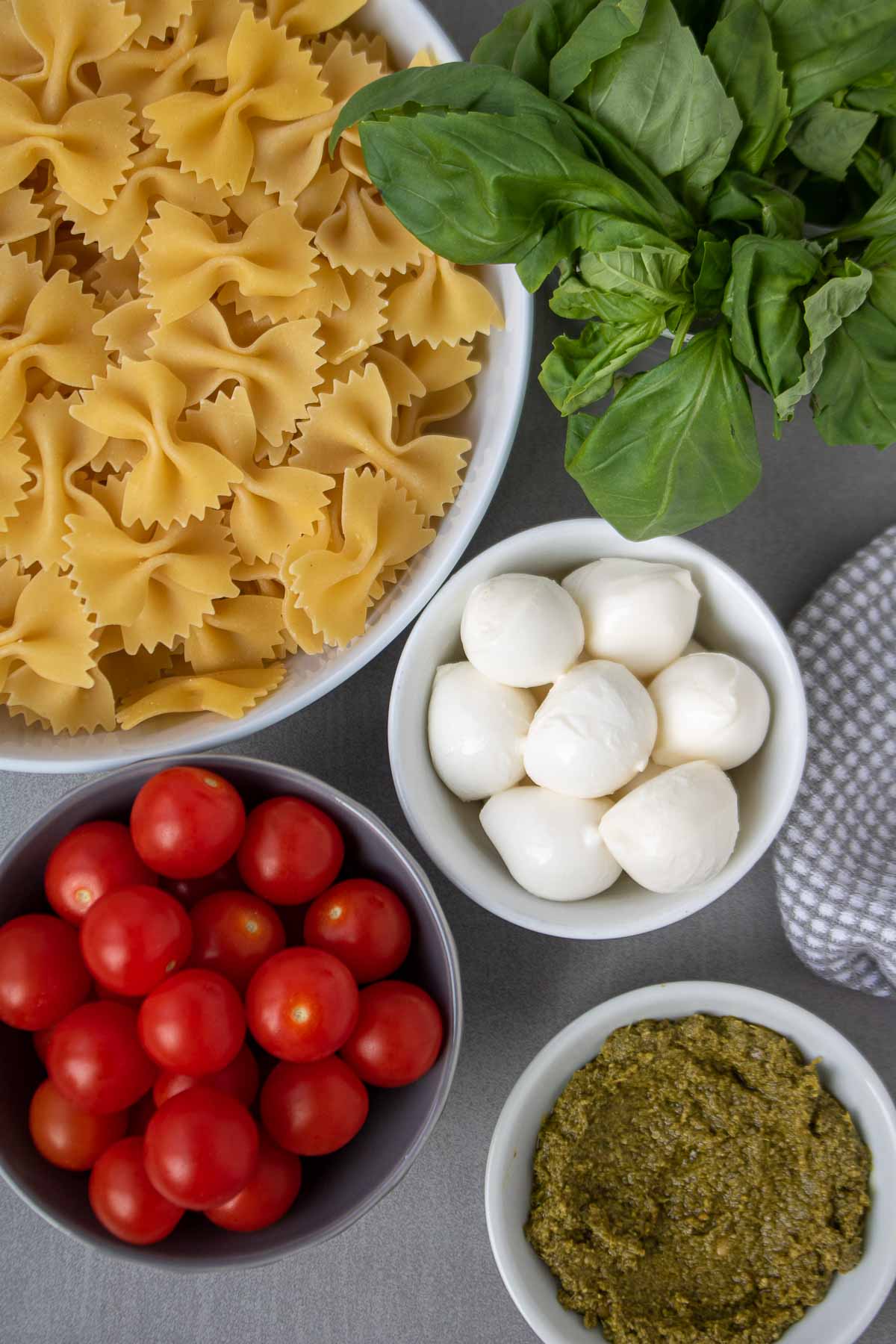 Ingredients for bow tie pasta salad: farfalle pasta, mozzarella cheese, tomatoes, basil, and pesto.