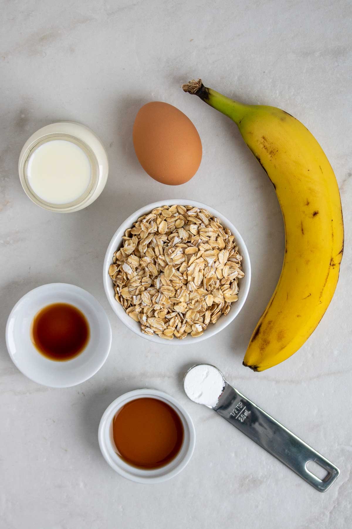Rolled oats, egg, banana, milk, vanilla extract, baking powder, and maple syrup on a white background