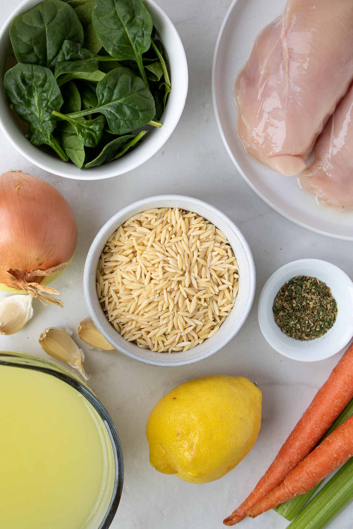 Ingredients for lemon orzo chicken soup; chicken breasts, orzo, spinach, onion, garlic, carrot, celery, basil, dill, oregano, parsley, salt, pepper, lemon, and chicken broth