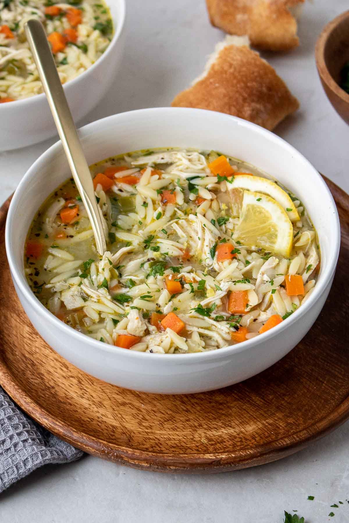 Chicken lemon orzo soup in a white bowl with a spoon, topped with lemon slices, and a side of crusty bread