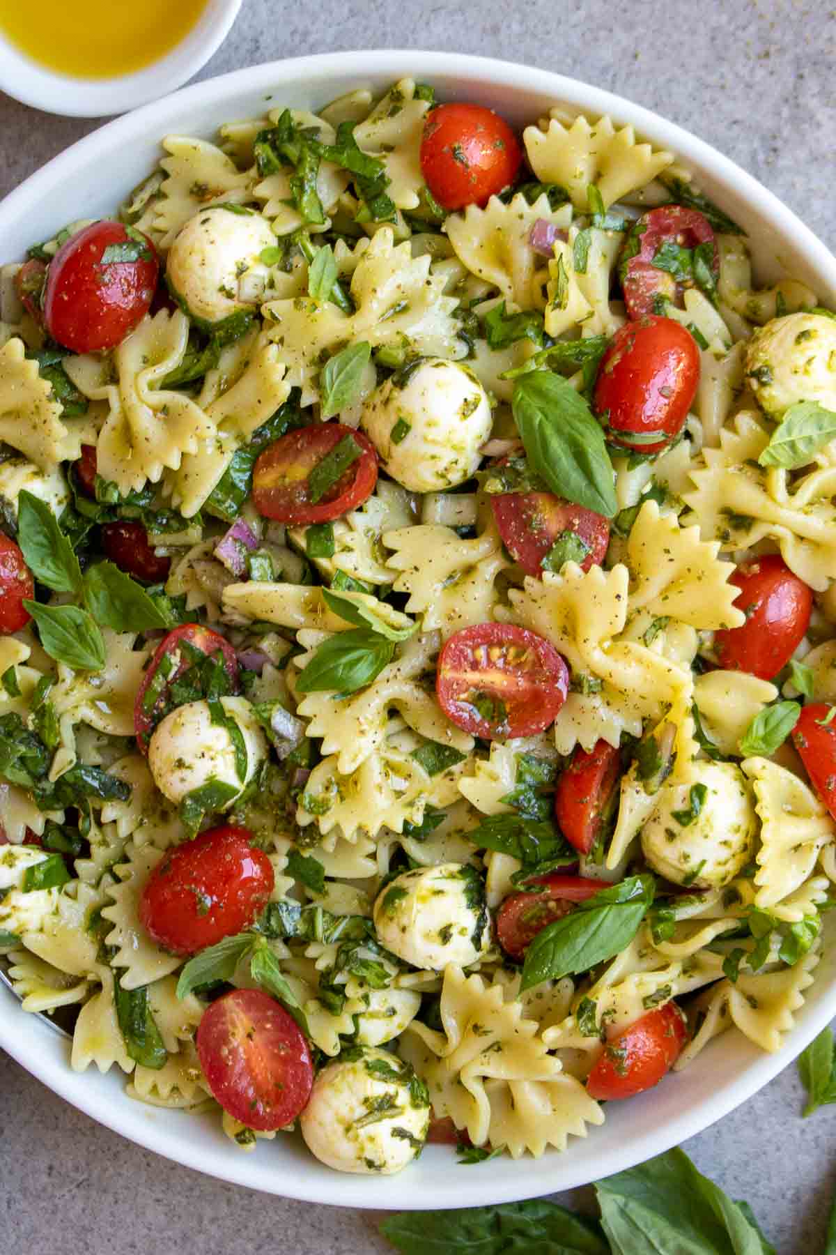A white bowl brimming with bow tie pasta salad adorned with tomatoes, spinach, basil, bocconcini cheese, pesto, olive oil, and a hint of lemon juice.