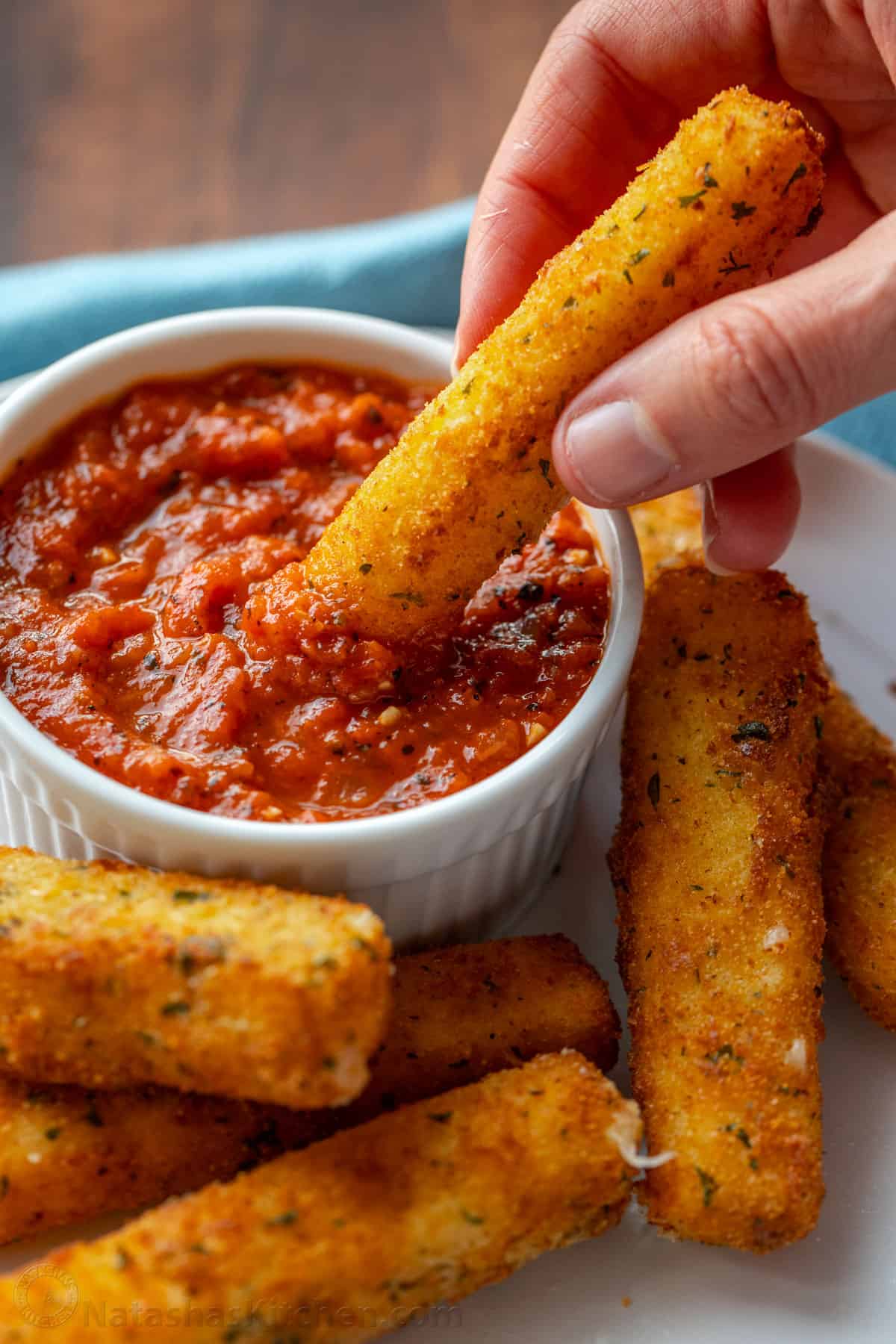 Une main trempe un bâtonnet de fromage mozzarella dans un bol de sauce marinara, à côté de plus de bâtonnets de fromage sur une assiette.